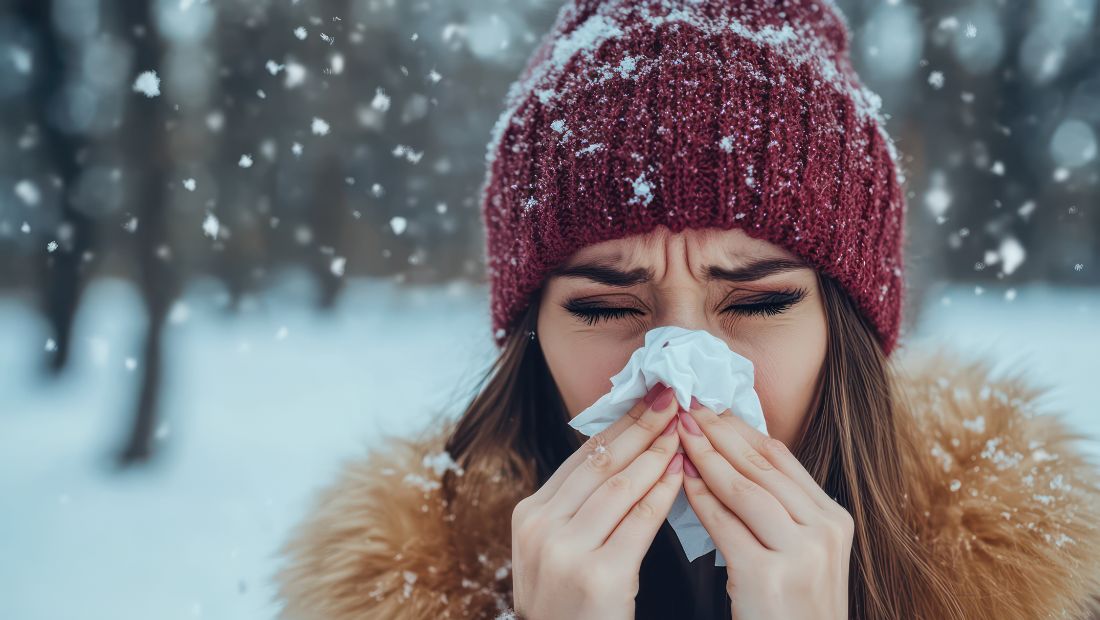 Osoba vonku v zimnom prostredí, oblečená v teplom oblečení, drží vreckovku pri nose a smrká.