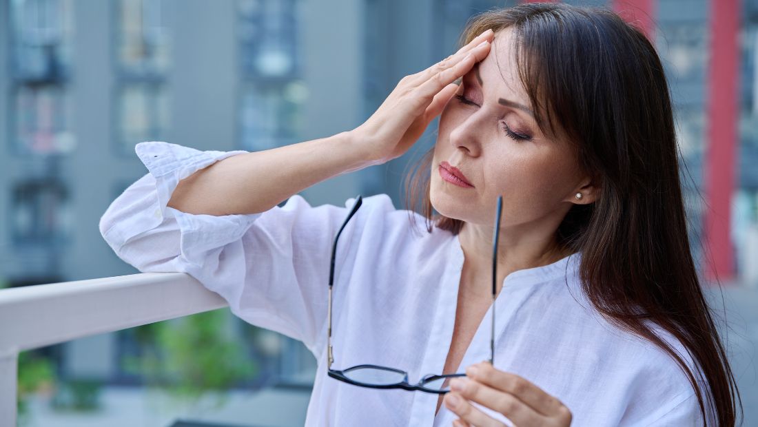 Žena prožívající nával horka při menopauze, drží v ruce brýle a chladí se na balkoně.